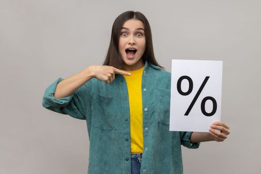 Amazed surprised woman holding and pointing at paper with percent sign inscription, looking at camera with open mouth, wearing casual style jacket. Indoor studio shot isolated on gray background.