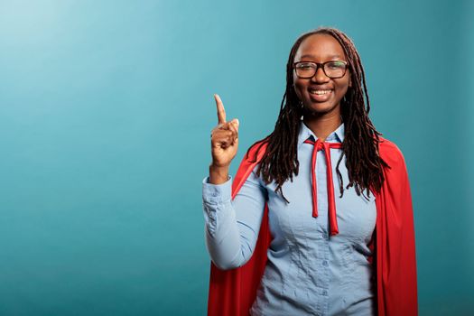 Optimistic happy superhero woman wearing red cape pointing finger up while standing on blue background. Proud and positive justice defender smiling at camera while gesturing fingers up. Studio shot