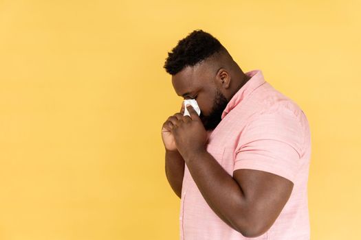 Side view portrait of man wearing pink shirt hiding face in hand with white handkerchief, crying, feeling stressed, worried facial expression. Indoor studio shot isolated on yellow background.