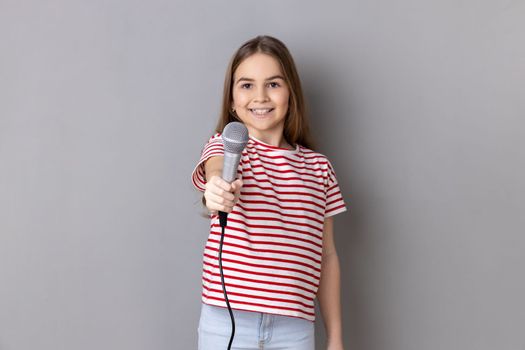 Portrait of cute little girl wearing striped T-shirt posing with microphone in hands, offers mic, asking questions, looking at camera. Indoor studio shot isolated on gray background.