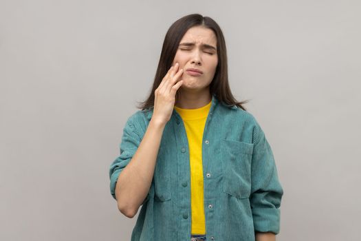 Unhealthy woman touching cheek, closing eyes with expression of terrible suffer from toothache, sensitive teeth, cavities, wearing casual style jacket. Indoor studio shot isolated on gray background.
