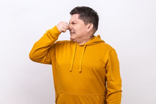 Middle aged man holding breath and grimacing in disgust, expressing repulsion to stink, fart gases, intolerable odor, wearing urban style hoodie. Indoor studio shot isolated on white background.