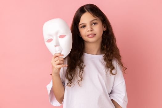Portrait of charming adorable dark haired little girl wearing white T-shirt holding white mask in her hand, wants to change personality. Indoor studio shot isolated on pink background.
