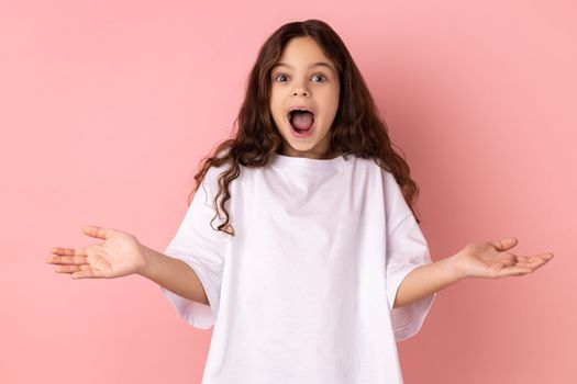 Wow, amazing. Amazed little girl wearing white T-shirt looking at camera with wide open mouth, expressing shock and excitement about great news. Indoor studio shot isolated on pink background.