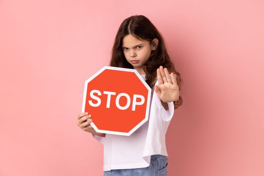 No, prohibited. Strict bossy little girl in T-shirt looking seriously at camera and showing stop gesture, holding road traffic sign as warning of ban. Indoor studio shot isolated on pink background.