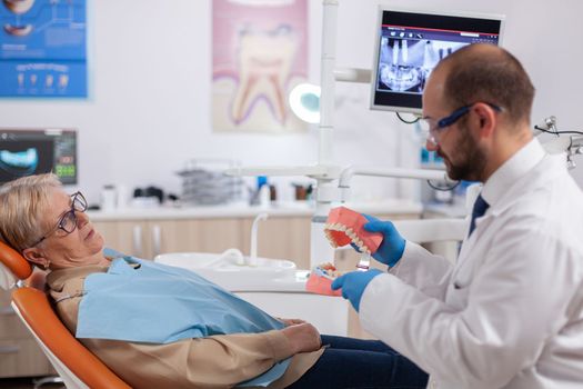 Dentist holding model of oral cavity with teeth during examination of senior patient. Medical teeth care taker discussing with senior woman about mouth hygiene.