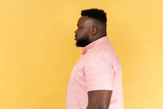 Portrait of serious strict young adult man wearing pink shirt standing and looking ahead with bossy facial expression, being in bad mood. Indoor studio shot isolated on yellow background.