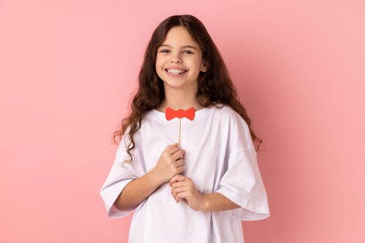 Portrait of satisfied little girl wearing white T-shirt holding funny paper bow tie, having fun playing game, wearing masquerade accessory. Indoor studio shot isolated on pink background.