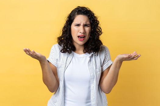 Portrait of unhappy stressed beautiful woman with dark wavy hair standing raised arms and asking, expressing negative emotions. Indoor studio shot isolated on yellow background.