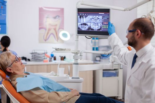 Stomatolog talking about teeh treatment to senior woman in dentist cabient. Medical teeth care taker pointing at patient radiography on screen sitting on chair.