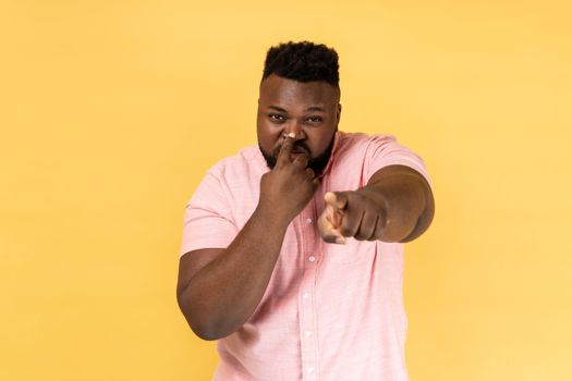 You are liar. Portrait of man wearing pink shirt standing with finger on her nose and showing lie gesture, pointing to camera. Indoor studio shot isolated on yellow background.