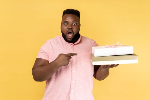 Portrait of amazed surprised shocked man wearing pink shirt holding and pointing at gift boxes, looking at camera with open mouth. Indoor studio shot isolated on yellow background.