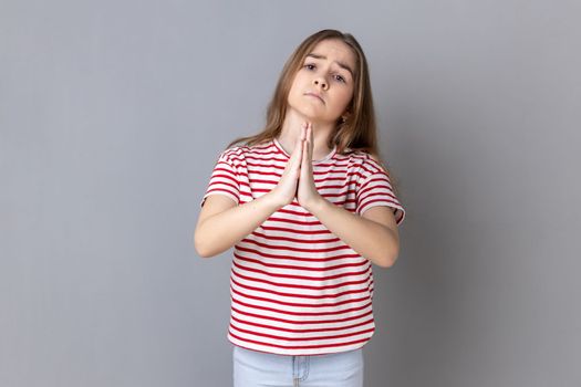 Please help me or forgive. Portrait of worry sad little girl wearing striped T-shirt standing with palm hands and looking at camera pleased pleading. Indoor studio shot isolated on gray background.