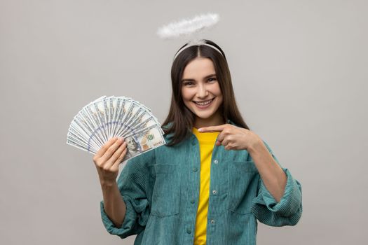 Cheerful dark haired woman with halo on head pointing finger on bunch of dollar banknotes and smiling, profit, wearing casual style jacket. Indoor studio shot isolated on gray background.