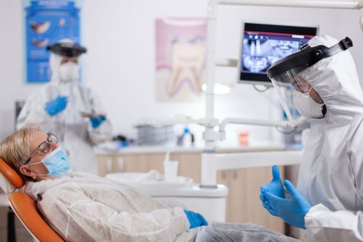 Dentist wearing safety gear against coronavirus talking about teeth treatment. Elderly woman in protective uniform during medical examination in dental clinic.