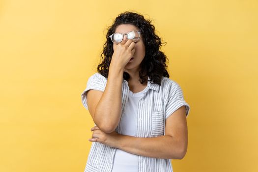 Portrait of tired exhausted attractive young adult woman with dark wavy hair standing with raised eyeglasses, feels pain in eyes. Indoor studio shot isolated on yellow background.