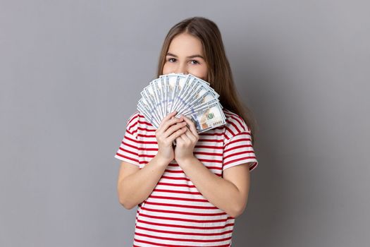 Portrait of charming little girl wearing striped T-shirt hiding half of face behind fan of hundred dollar bills, enjoying smell of money. Indoor studio shot isolated on gray background.