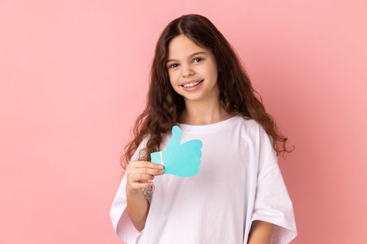Portrait of smiling delighted adorable little girl wearing white T-shirt holding and showing like or thumbs up paper shape sign. Indoor studio shot isolated on pink background.
