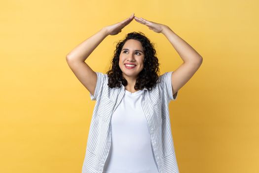 I'm in safety. Portrait of woman with dark wavy hair raising hands showing roof gesture and smiling contentedly, dreaming of house, looking away. Indoor studio shot isolated on yellow background.