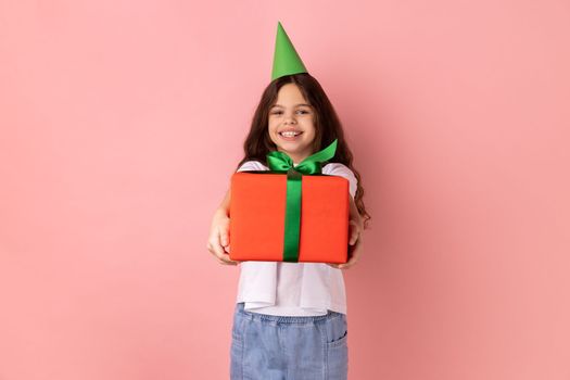 Portrait of little girl wearing white T-shirt with festive mood standing in party cone and giving present box, celebrating friend's birthday. Indoor studio shot isolated on pink background.