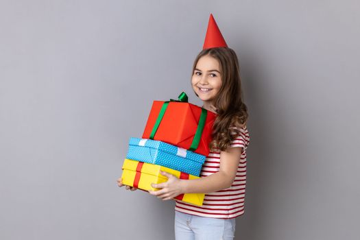 Portrait of delighted charming little girl wearing striped T-shirt and party cone, embracing stack of presents, celebrating birthday. Indoor studio shot isolated on gray background.