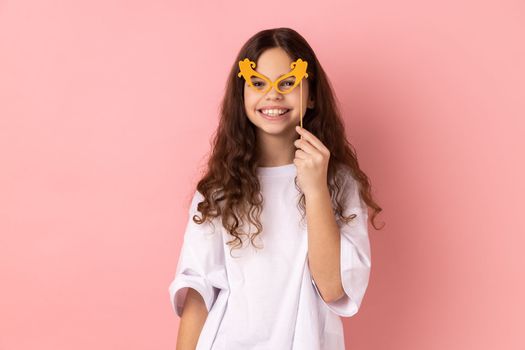 Portrait of cute happy little girl wearing white T-shirt smiling and holding paper glasses on stick, using party props on holiday, having fun. Indoor studio shot isolated on pink background.