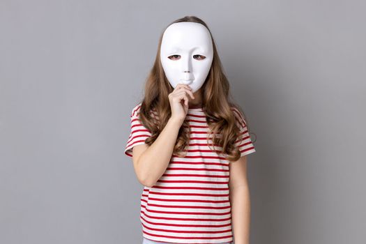 Portrait of anonymous unknown little girl wearing striped T-shirt covering her face with white mask, hiding her real personality, anonymity. Indoor studio shot isolated on gray background.