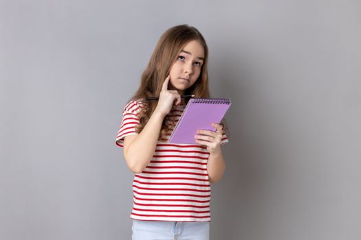 Portrait of thoughtful pensive little girl wearing striped T-shirt writing in organizer, making to do list, drawing or taking notes. Indoor studio shot isolated on gray background.