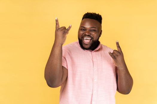 Portrait of disobedient man wearing pink shirt showing rock and roll gesture and excitedly screaming, having fun, enjoying heavy music. Indoor studio shot isolated on yellow background.