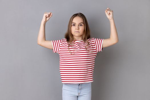 Strong dark haired little girl wearing striped T-shirt standing and raising arms, showing her strength and looking at camera with serious expression. Indoor studio shot isolated on gray background.