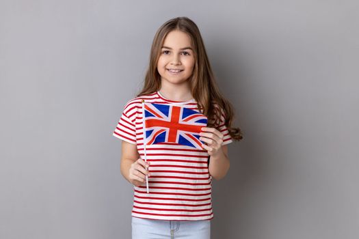 British flag. Portrait of charming smiling little girl wearing striped T-shirt holding flag of United Kingdom looking at camera with positive expression. Indoor studio shot isolated on gray background