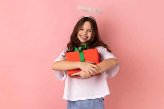 Portrait of little girl wearing white T-shirt and angelic halo embracing wrapped box, enjoying birthday gift, satisfied with long-awaited present. Indoor studio shot isolated on pink background.