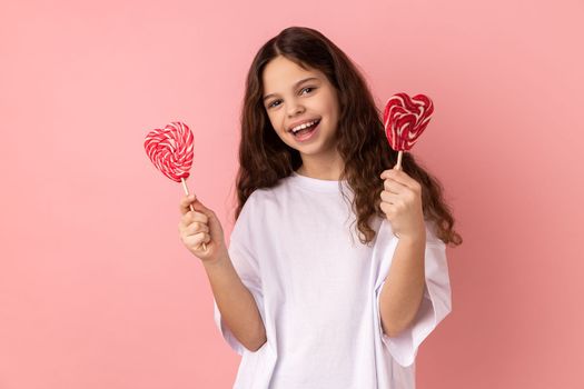 Portrait of satisfied little girl wearing white T-shirt holding two heart shape lollypops, looking at camera with happy expression. Indoor studio shot isolated on pink background.
