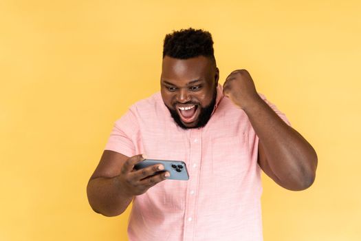 Portrait of man wearing pink shirt standing, using smartphone and playing mobile game with excited positive face, happy to win, clenched fist. Indoor studio shot isolated on yellow background.
