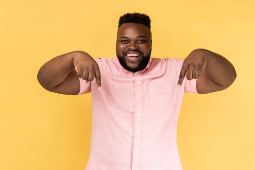 Look, advertise below. Portrait of delighted optimistic man in pink shirt pointing down place for commercial idea, looking at camera with toothy smile. Indoor studio shot isolated on yellow background