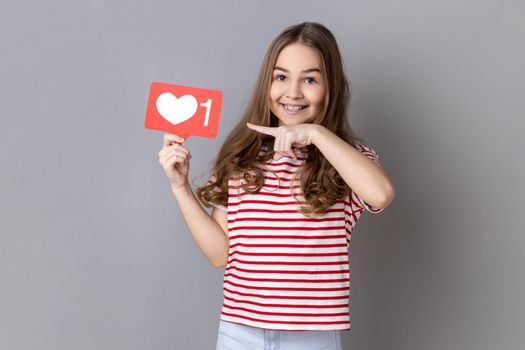 Portrait of adorable smiling little girl wearing striped T-shirt pointing at heart like icon, recommending to click on social media button. Indoor studio shot isolated on gray background.