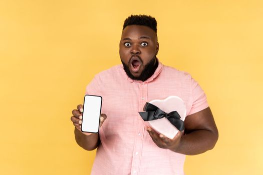 Portrait of surprised shocked bearded man wearing pink shirt holding heart shaped present box and showing smart phone with empty screen. Indoor studio shot isolated on yellow background.