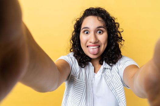 Portrait of funny positive woman with dark wavy hair making point of view photo or broadcasting livestream, showing tongue out, POV. Indoor studio shot isolated on yellow background.