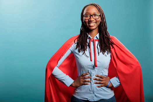 Proud and happy young superhero woman posing brave for camera while standing on blue background. Strong and selfless justice defender wearing mighty hero red cloak while standing with hands on hip.
