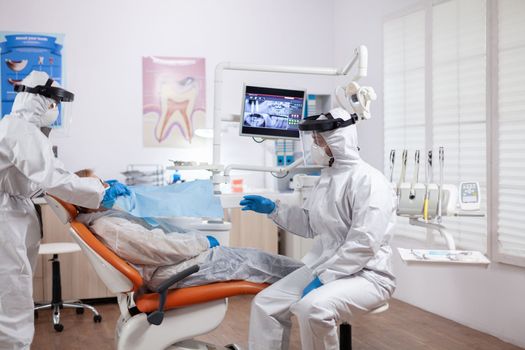 Dentist nurse taking patient temperature wearing hazmat suit using digital thermometer. Elderly woman in protective uniform during medical examination in dental clinic.