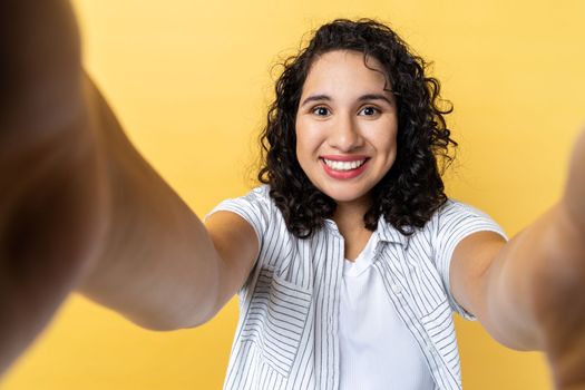 Portrait of charming cheerful woman blogger with dark wavy hair making point of view phone, looking with toothy smile, expressing happiness. Indoor studio shot isolated on yellow background.