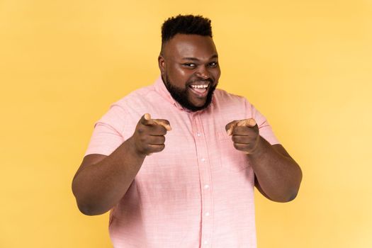Portrait of cheerful man wearing pink shirt pointing to camera and looking with charming toothy smile, indicating finger, making choice. Indoor studio shot isolated on yellow background.