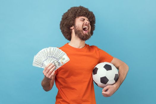 Excited man with Afro hairstyle wearing orange T-shirt holding soccer ball and hundred dollar bills, screaming with happiness, betting and winning. Indoor studio shot isolated on blue background.