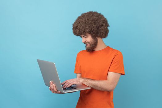 Portrait of man with Afro hairstyle in orange T-shirt holds laptop in hand and typing, blogger making posts in social networks, chatting with followers. Indoor studio shot isolated on blue background.