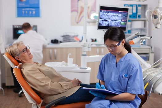 Assistant in dentist cabinet during elderly woman oral hygiene check up sitting on chair. Senior woman talking with medical nurse in stomatology office about teeth problem.