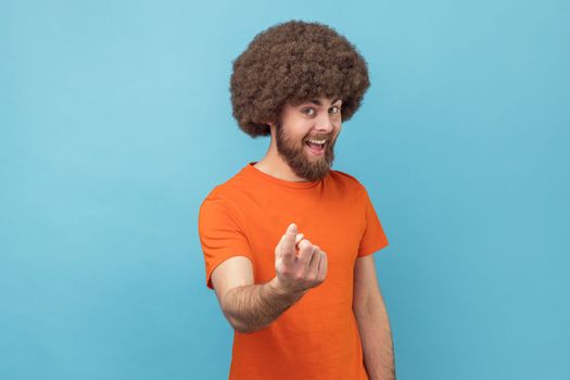 Come here. Portrait of satisfied smiling man with Afro hairstyle wearing orange T-shirt calling with finger gesture, inviting to come in. Indoor studio shot isolated on blue background.