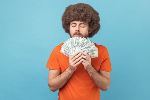 Portrait of man with Afro hairstyle wearing orange T-shirt smelling dollar banknotes with pleasure expression, enjoying big money profit and rich life. Indoor studio shot isolated on blue background.