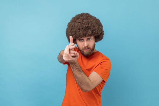 Portrait of serious man with Afro hairstyle wearing orange T-shirt holding fingers pretending holding gun, aiming enemy, self-defense. Indoor studio shot isolated on blue background.
