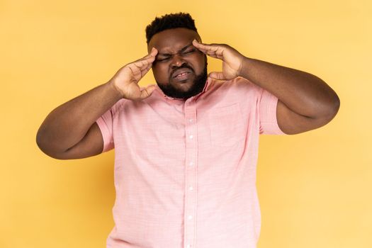 Portrait of unhealthy sick young adult man wearing pink shirt massaging temples, suffering from headache, migraine, frowning his face. Indoor studio shot isolated on yellow background.