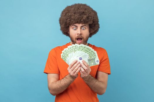 Portrait of shocked amazed man with Afro hairstyle wearing orange T-shirt holding bunch of euro banknotes, looking at money with open money. Indoor studio shot isolated on blue background.